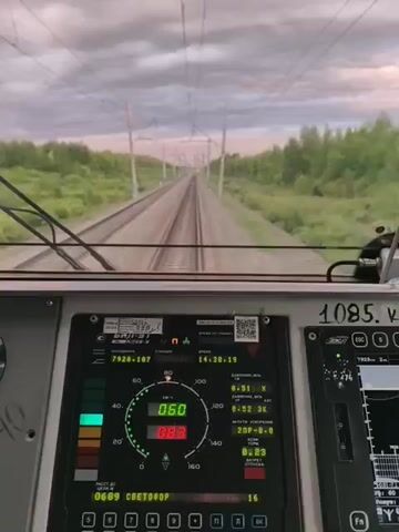 The video shows the interior of a train's cabin, looking out through the front windshield. The tracks of a railway line extend into the distance, flanked by lush green trees. Overhead, power lines crisscross the sky, which is filled with dramatic, cloud-filled twilight.

The central focus of the video is the train's control panel. A large digital display shows various readings, including speed, time, and other operational data. A speedometer indicates a speed of 60 km/h, with a red digital readout below showing 083. Another smaller display to the right shows a graph, likely representing speed or other operational parameters. On the upper right of the panel, the number "1085." is visible, along with the Cyrillic abbreviation "кр.".

The lighting suggests it is either early morning or late evening, with the soft, diffused light painting the sky in hues of pink and grey. The weather appears to be cloudy, contributing to the dramatic sky. The overall impression is one of a journey through a serene, natural landscape, viewed from the functional and high-tech environment of a train's cockpit. The presence of Cyrillic text on the control panel and the mention of "Tur, Russia" provide a clear geographical context.