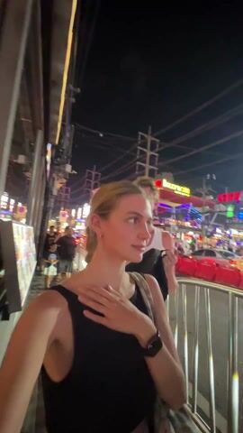 A woman is standing on a busy street at night. In the background is the entrance to a building that says "Bollywood". A car drives by with a flatbed on top that is showing an ad for Championship Fight Night, which is on Sunday the 5th. There are two shirtless men wearing gloves on the flatbed.