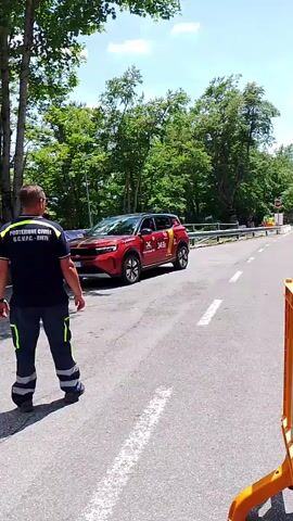 A person wearing a navy blue shirt with yellow accents and black pants is standing on the left side of a paved road. The shirt has text in yellow, and the pants have yellow reflective stripes. The person is facing away from the camera and towards the road. A red SUV is parked on the left side of the road behind the person. A white car is driving on the right side of the road. Another person is standing further down the road on the right, with their arms outstretched. There are trees and foliage on both sides of the road. An orange construction barrier is visible in the foreground on the right. The sky is blue with some clouds. The overall impression is that of a road closure or traffic management situation, possibly related to an event or construction.
