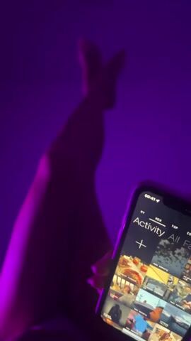 The video shows a person lying down with their legs crossed in the air, illuminated by a purple light.  In the foreground, the person is holding a smartphone displaying an activity feed or a similar app interface with various images and icons. The time displayed on the phone is 00:41. The background is a solid, dark purple.