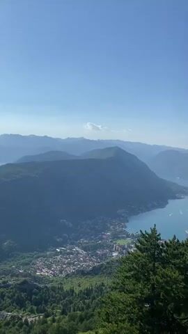 This video captures a sweeping panoramic view of a bay surrounded by mountains. The clear blue sky stretches across the top of the frame. In the mid-ground, a body of turquoise water, likely a bay or fjord, is dotted with several small white boats, indicating moderate maritime activity. Along the edge of the water, a coastal town with numerous buildings is visible, nestled at the foot of the imposing, verdant mountains. The mountains themselves are densely covered in trees, with some rocky outcrops visible. In the foreground, the tops of two large, dark green pine trees partially obscure the view, adding a sense of depth to the scene. The overall impression is one of a beautiful, sunny day in a mountainous coastal region.