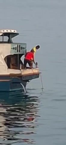 The video shows a wide shot of a boat with two men on the stern. One man, wearing a red shirt, is kneeling and appears to be operating a winch or pulley system. The other man, wearing a yellow shirt, is standing and looking down over the side of the boat, possibly observing the operation or preparing to dive. The boat is in calm, blue water. The camera is positioned at eye level with the boat.