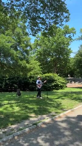 This is a video of a man playing an acoustic guitar and singing in a park. The man is wearing a hat and a vest over a white shirt and dark pants. He is standing in a grassy area, with a microphone stand in front of him. A black chair is placed to his left. Tall green trees with full canopies dominate the background, casting dappled shadows on the ground. The sky is a clear, bright blue, suggesting a sunny day. In the distance, other park-goers can be seen, including some children playing. The overall setting appears to be a well-maintained public park in New York City, possibly during the daytime given the bright sunlight and clear sky. The mood seems peaceful and enjoyable, with the musician providing entertainment for those in the park. There is no visible text in the video.