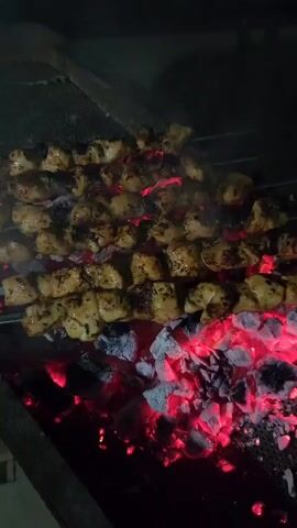 The video shows chicken skewers being grilled over hot coals. The skewers are arranged on a metal grill. The coals are glowing red and white. Smoke is rising from the grill. The background is dark and out of focus.