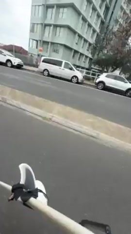 The video opens with a street scene with a tall, modern building in the background under a cloudy sky. A white SUV and a white van are parked on the side of the road. The camera then pans to the left, showing a row of cars parked along the street, including a dark grey sedan. A person wearing a black helmet and riding a black scooter then comes into view. The camera's perspective appears to be from the point of view of the scooter rider. The rider turns the scooter and appears to drop the camera as the view goes dark. The camera then abruptly shifts, facing upward toward the cloudy sky and the building.