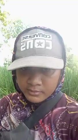 A person is shown from the chest up, facing the camera. They are wearing a grey baseball cap with white lettering that reads "CONVERSE" and a star logo. The person also has on a hooded shirt with a patterned design in shades of red, purple, and white, and a black strap is visible across their chest. The background consists of tall, green grass and some blurred trees. The lighting suggests it is daytime.