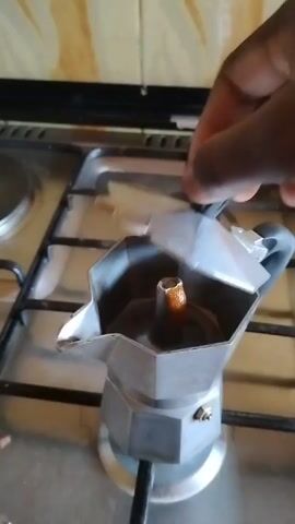 The video shows a person opening a Moka pot sitting on a stainless steel gas stovetop. The Moka pot is made of grey metal and has an octagonal shape. The pot's top compartment is open, revealing the funnel containing used coffee grounds and a small amount of dark liquid. The lid is held open by the person's hand, which is dark-skinned. The stovetop has multiple burners, some with metal covers, and a black backsplash. There's a small metal hook and what appears to be a piece of food debris on the stovetop. The video seems to be taken in a kitchen.