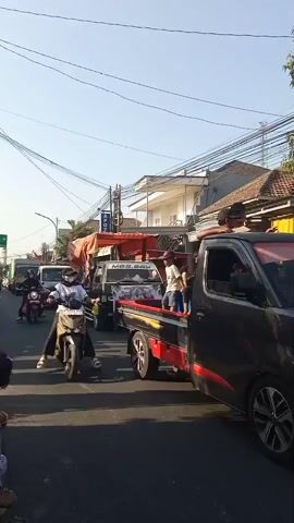 The video shows a street scene with several vehicles and people. In the foreground, a person is riding a beige scooter. The scooter has a black seat and front fender. The rider is wearing a black jacket and a white shirt with a logo. Behind the scooter, a black pickup truck is visible. The truck has red trim along the side and a black bed. Several people are standing in the bed of the truck. One person, wearing a white shirt and blue shorts, is holding onto a pole. Another person, wearing a dark shirt and shorts, is also in the truck bed. Further down the street, a white truck is parked. The truck has an orange tarp covering its load. A group of people are gathered around the back of the truck. The street is lined with buildings and power lines are visible overhead. The sky is clear and the sun is shining.