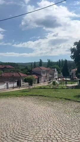 This video captures a serene village scene in Zelenikovo, Bulgaria. The foreground features a cobblestone street, leading the eye towards a well-maintained grassy area with a playground. Two figures, likely children, are visible on the playground, suggesting a time of leisure and outdoor activity. Beyond the playground, a row of traditional houses with red-tiled roofs lines the street, nestled against a backdrop of lush green hills and trees. The architecture hints at a rural European setting.

The sky is a bright blue, dotted with fluffy white clouds, indicating pleasant weather, likely mid-morning or afternoon given the strong shadows cast by the trees and a prominent utility pole. The overall mood is peaceful and idyllic, reflecting the quiet charm of village life. The scene is bathed in natural sunlight, highlighting the vibrant green of the grass and the rustic textures of the cobblestones and rooftops. There are no discernible signs or text visible within the frame. The video provides a snapshot of a typical day in this Bulgarian village, emphasizing its natural beauty and community spaces.