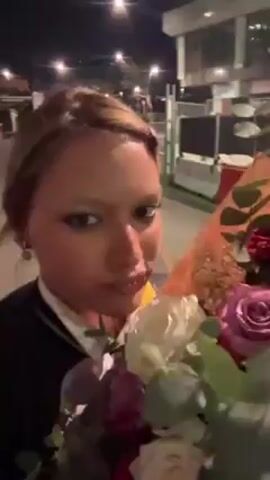 A woman with light brown hair, wearing a dark blazer with a white collar, is holding a bouquet of roses in front of her face. The roses are various colors including red, purple, and white. The woman is looking directly at the camera and smiling slightly. In the background, there's a building lit up at night, possibly an event venue, with other indistinct figures visible. The setting appears to be outdoors, with streetlights contributing to the illumination.