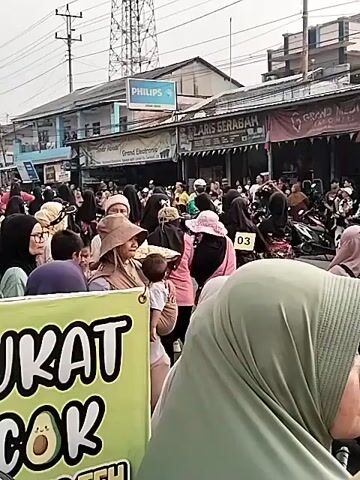 The video shows a crowded street scene with many people, most of whom are wearing headscarves. There are shops and stalls lining the street, with signs visible for businesses like "Grand Electronic" and "Philips." In the foreground, a large green banner with white lettering is partially visible, with the words "UKAT COK" and an image of an avocado. Several motorcycles are parked along the street, and some people appear to be interacting with vendors or browsing the shops. The overall atmosphere seems to be that of a bustling market or gathering.