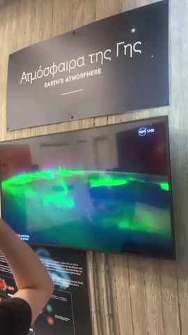 The video displays a close-up view of a television screen showing an aurora borealis effect. The aurora is depicted with vibrant green and purple hues, undulating across the screen. Above the television, a sign reads "Ατμόσφαιρα της Γης EARTH'S ATMOSPHERE" in Greek and English. Below the television, there is a partial view of another informational display, with some Greek text visible. A person's arm and shoulder are partially visible in the lower left corner of the frame, extending into the scene from off-camera. The television screen is reflective, showing a blurred image of the surrounding environment, including what appear to be doorways or openings in a wall. The background wall behind the sign has a textured, wood-like finish.