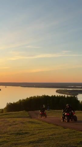 Two individuals are riding ATVs on a dirt path atop a grassy hill, with a scenic sunset view of a wide river and islands in the background. A group of people stand near the edge of the hill, possibly enjoying the view. Another person walks into the frame on the right side of the video. The sky is a blend of blue and orange hues, typical of a sunset. The hill is covered in green grass and bordered by trees.