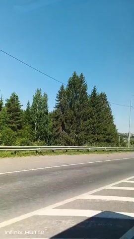 A video shows a road with a bright blue sky above and a line of trees on the left side. The road has white markings on it. In the distance, there are some houses and more trees. There are also some utility poles and wires visible. The video appears to have been filmed from a moving vehicle. The lower left corner of the video displays the text "Infinix HOT 50".