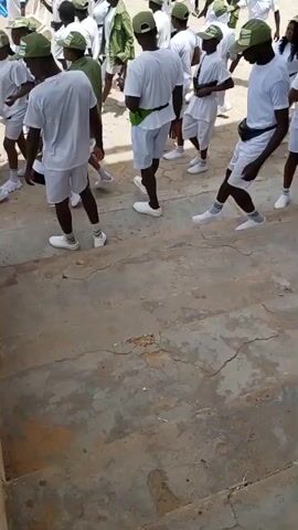 This media file captures a group of young people, likely participants in a program or event, dressed in matching white t-shirts and shorts, with green caps. The setting appears to be an outdoor area with concrete stairs and a rough, possibly dusty ground. In the background, a larger crowd of individuals in similar attire is visible, along with some green structures and a white tent or canopy.

The individuals in the foreground are mostly facing away from the camera, with some engaged in movement, suggesting they are either marching, dancing, or participating in some form of organized activity. One person in the front right is looking at a mobile phone. The overall atmosphere seems to be one of a communal gathering, possibly a training camp or orientation program.

The clothing suggests a uniform, indicating a specific group or event. The time of day appears to be daytime, judging by the lighting, and the weather seems to be bright and sunny. The concrete stairs and the textured ground suggest an outdoor public space, potentially a training ground or assembly area. No visible text is present in the media file. The individuals' postures and the group formation hint at a structured activity.