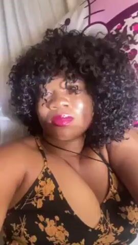 A woman with dark skin, black curly hair and shiny red lipstick lies on her back on a bed with a Hello Kitty pillow visible in the background. She is wearing a black halter top with yellow floral patterns. She sticks out her tongue and puckers her lips.