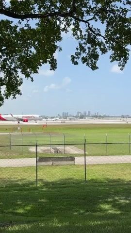 A video shows an Avianca airplane on a runway on a sunny day. The plane is white with a red tail. In the background, a city skyline is visible. The foreground shows a green grassy area with a chain-link fence. Trees partially obscure the view. The plane appears to be taxiing or preparing for takeoff.