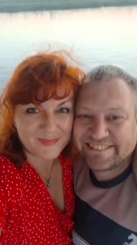 This is a selfie-style video of a Caucasian couple, appearing to be in their 40s or 50s, standing close together outdoors. The woman has vibrant red hair and is wearing a red polka-dot dress. The man has short, graying hair and stubble. They are both smiling and looking at the camera. The background suggests they are near water, possibly a lake or river, with a hazy horizon. The lighting is soft, indicating it might be daytime, perhaps late afternoon or early morning. There is no visible text in the video. The overall mood is cheerful and intimate, suggesting a personal moment captured during an outing.