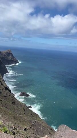 A high-angle shot displays a vast expanse of the ocean, transitioning in color from turquoise near the rocky coastline to a deeper blue in the distance. The sky is partly cloudy with patches of blue visible. The camera appears to be positioned on the edge of a cliff or high point, with vegetation visible on the rocky foreground. The sea waves are crashing against the rocks along the coastline.