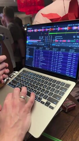 The video shows a DJ performing in a crowded bar or club. The DJ is operating a laptop and mixing board. The laptop displays a DJ software interface. Several people are in the background, some dancing and others talking. One person holds a small camera and records the DJ and the crowd. The lighting is dim, typical of a nightlife setting. A decorative red object hangs from the ceiling.