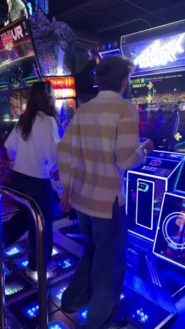Two people are playing a rhythm game in an arcade. They are standing on a dance pad with illuminated squares that correspond to directional arrows on the screen. The person on the right, a male wearing a striped shirt and jeans, is actively stepping on the pads, while the person on the left, a female wearing a white shirt and dark pants, is also engaged with the game. The arcade environment is dimly lit, with bright neon lights emanating from the game machines, creating a vibrant and energetic atmosphere. The screens display colorful graphics and scrolling notes, indicating the gameplay in progress. A metal railing is visible in the foreground, separating the players from the viewer's perspective.