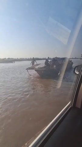 The video shows a river scene from inside a vehicle. A flat-bottomed boat is visible in the water, carrying several people and at least one motorcycle. One person is standing at the front of the boat, possibly using a pole to navigate. The water is murky, and the sky is clear with a bright sun. The landscape appears to be flat, with some vegetation visible in the distance. The boat is moving across the river.