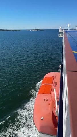 This video shows a view from the side of a large ship, likely a ferry, moving through calm, blue water on a clear, sunny day. The camera is positioned looking forward and slightly down, capturing the wake of the ship. In the foreground on the right is the side of the ship with a white railing and a reddish-brown top edge. Attached to the side of the ship is an orange lifeboat, labeled with the number 2 and the IMO number 9663003. In the distance, across the water, is a landmass with some structures, possibly an island or a coastline. The water is a deep blue, with white foam from the ship's wake. In the final frame, a man in a light-colored shirt is visible standing on the deck of the ship, leaning against the railing, looking out at the sea. Another person is partially visible behind him. The sky is a clear, bright blue.
