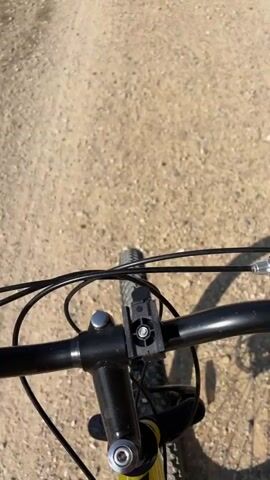 The video shows a first-person perspective of someone riding a bicycle on a dirt path. The rider is looking down at the handlebars and the path ahead. The path is surrounded by green foliage on the right side. The sun is shining, casting shadows. At 00:03, another cyclist, a young girl on a yellow bicycle, is seen riding ahead on the same path. She is wearing a white t-shirt, patterned shorts, and a pink hat. The camera continues to follow her for a few seconds before the video ends.