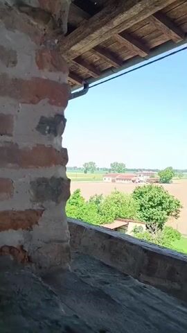 The video shows a view of a flooded field and buildings in the distance, framed by an old brick structure in the foreground. The brick structure appears to be part of a building with a wooden roof overhang. The field in the distance is covered with water, and several trees and buildings are visible. The sky is clear and blue.