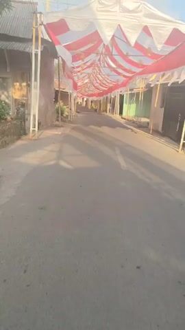 The video shows a street lined with buildings and decorated with a canopy of red and white striped fabric. The canopy extends down the street, supported by poles on either side. Ornate lanterns hang from the canopy. The street is paved and has a white line marking the center. Shadows of trees and possibly a person on a motorcycle are cast on the road. Some motorbikes are parked along the side of the street. The environment appears to be a village or a small town.