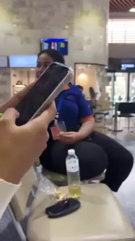 A woman in a white sweater holds a smartphone and looks at the screen, then looks at a man in a blue and orange polo shirt. The man is sitting near another person wearing the same type of shirt. The woman with the phone is seated, and there is a water bottle, a pen, and what appears to be a set of car keys on the chair next to her. The scene appears to be taking place indoors, possibly in an airport waiting area, with tile flooring and a high ceiling. A digital display and service counters are visible in the background.