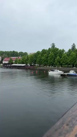 The video opens with a wide shot of a calm body of water with a cloudy sky overhead. A distant shoreline is visible with green trees and buildings, some with red roofs. Several boats are docked or anchored near the shore, including a white motorboat with a cabin on the right side of the frame. Red buoys are scattered on the water. The camera pans slowly to the left, revealing more of the shoreline with additional buildings, green trees, and parked boats. Rain is visibly falling, creating small ripples on the water's surface. A faint, low-pitched sound of traffic and distant voices can be heard throughout the video. The video ends as the camera continues panning left, showing more of the distant shoreline.