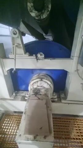 The video shows a static shot of a piece of industrial machinery. The machine is primarily white and blue, with a large blue disc visible between the structural supports. A dirty, cylindrical component is positioned at the bottom center of the frame, partly obscured by a brown piece of material. The machine sits on a yellow, gridded surface. The environment appears to be dimly lit.