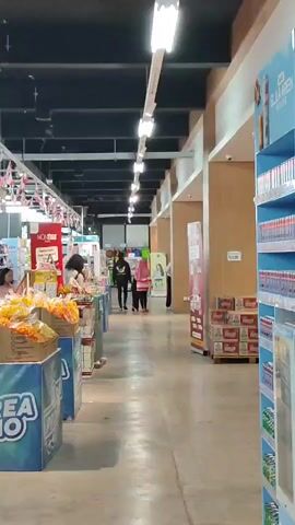 The video shows an interior view of a supermarket aisle. Bright fluorescent lights illuminate the scene from the ceiling. On the left side of the aisle, there are shelves stocked with various products, including what appear to be snacks in bags. In the foreground, large blue displays with the text "AREA PROMO" are visible. Further down the aisle, two individuals, one wearing a black hoodie and the other a pink hijab, are walking away from the camera. Another person, wearing a white shirt and dark pants, is interacting with products on a shelf. On the right side of the aisle, stacked boxes of beverages are visible, along with a blue shelf unit containing numerous bottles. The floor is a polished concrete. The overall impression is a typical retail environment.