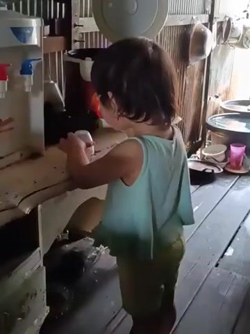 A young child, possibly a toddler, stands at a water dispenser. The child is wearing a light blue tank top and shorts. Their hands are on the dispenser, and they appear to be pressing buttons or operating a lever. The water dispenser has a red and blue knob, suggesting hot and cold water. The background shows a rustic environment with wooden walls and floor, and kitchen utensils hanging on the wall. The scene suggests a domestic setting, possibly a kitchen or utility area. The child's actions indicate an interaction with the water dispenser, possibly trying to get water.