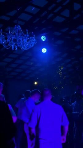 This is a vertical video shot from a low angle inside what appears to be a dimly lit venue with a dark, grid-like ceiling structure. A large, ornate crystal chandelier hangs from the ceiling on the left side of the frame, illuminated with blue light. Above and to the right of the chandelier, two round blue stage lights are visible, emitting a bright blue glow. The space is filled with a crowd of people, mostly seen from behind, with some individuals dancing and socializing. Their silhouettes are dark against the blue lighting. The overall atmosphere is that of a party or event, with the blue lighting creating a club-like ambiance. Green foliage is visible on the right side of the frame, adding to the decorative elements of the venue. The video captures a brief moment of activity within this setting.