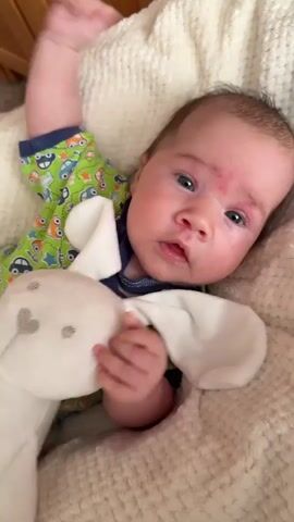 A close-up shot shows a baby lying on its back on a fluffy cream-colored blanket. The baby is wearing a green onesie with a colorful car print. In the baby's left hand, it holds a white stuffed rabbit toy with long floppy ears. The baby's head is tilted slightly to the right, and its eyes are open, looking towards the camera. A red mark is visible on the baby's forehead. The lighting is bright, suggesting it is daytime. The background is softly blurred, with a hint of wood grain visible, possibly from furniture. The camera angle is from above, looking down at the baby.