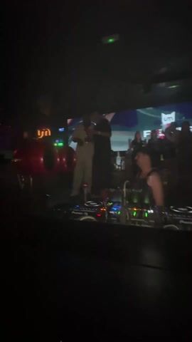 The video shows scenes from inside a crowded nightclub. The initial shot is from behind a DJ console with people on stage. The video then pans to the crowd, showing various people dancing and singing along to the music. The lighting is dark with bright flashes of light.