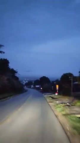A POV shot of a dark paved road at dusk. The road curves gently to the left and then to the right, leading toward a distant town visible through the mist. Trees and bushes line the sides of the road, with power lines and poles visible on the left. A few lights from buildings can be seen in the distance. The sky is a deep blue, indicating fading daylight. The headlights of the vehicle are visible, illuminating the road ahead.