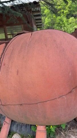 The video shows two large, reddish-brown clay pots in a yard. The first pot is rotund and sitting on small, leg-like supports. It has a large, circular opening and some wire wrapped around its body to hold it together. The second pot is taller and more egg-shaped, with a grid of wire surrounding it. Both pots appear to be outside, near a small wooden building and surrounded by green foliage and grass.  A red hose lies near the egg-shaped pot.
