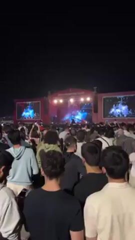 Here is a concise description of the scene in the video:

The video shows a concert at night. There is a large crowd gathered facing a stage with a band performing under bright lights. Two large screens flank the stage, displaying the performance. The people in the crowd are facing the stage, and some are holding up their phones to record the event.
