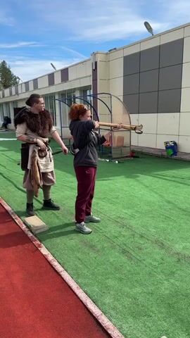 The video shows two people outdoors, one in medieval clothing standing next to the other, who is holding a wooden crossbow and aiming at a target. The target is off-screen to the right of the frame. The woman pulls the trigger and the projectile is released. The camera pans slightly to follow the trajectory of the projectile, which appears to fall short of its target. The camera then returns to the woman, who is looking toward the target area. The setting appears to be on artificial turf next to a light-colored building with a red running track on the far left of the frame. The sky is partially cloudy.