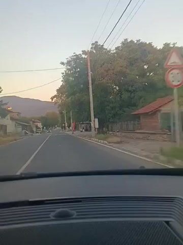 A POV shot from a car driving down a paved road. The sky is a light orange and the sun is low, suggesting it is either sunrise or sunset. On the left side of the road, there are some buildings and a mountain range in the distance. On the right side of the road, there is a large tree, a utility pole with wires, a bus stop, and what appears to be a train track running parallel to the road. There are several cars on the road, both moving and parked. A speed limit sign of 30 is visible on the right side of the road. The interior of the car is visible in the foreground, with the dashboard and windshield.