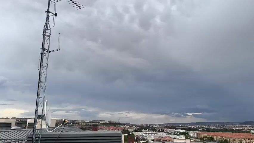 The video provides an elevated, static view of an urban or suburban landscape under a heavily overcast sky. In the foreground, a rooftop with a tall metal antenna mast, equipped with multiple television aerials and partially visible satellite dishes, is prominent on the left.

The mid-ground showcases a sprawling town or city, composed of numerous buildings, some featuring reddish-orange tiled roofs characteristic of the region. Distant hills or low mountains are visible on the horizon in the background.

The sky is dominated by dark, tumultuous grey clouds, indicative of an impending or ongoing weather event. A distinct curtain of rain is visible falling in the distance under the darkest cloud mass, suggesting localized precipitation. The overall lighting is dim due to the dense cloud cover, hinting at either late afternoon or early evening. The camera briefly pans slightly to the right at the beginning before settling. No people or visible text are present in the video. The location context is Arroyo de la Encomienda, Spain.