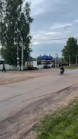 The video shows a rural scene, likely a village. Two young men are riding a small motorcycle down a paved road. The rider is wearing a dark jacket and pants, and the passenger is wearing a light-colored hooded jacket. They are riding side-by-side on the motorcycle. In the background, there are houses, trees, and power lines. The sky is cloudy, with some hints of pink and orange from what appears to be a sunset. Several other people are visible in the background, walking and standing near buildings. One person is walking on the side of the road, and another is near a parked truck. The overall impression is of a casual, everyday scene in a rural setting.