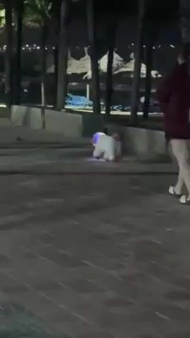 At night, a girl with red hair wearing a dark red jacket and white leggings stands on a brick promenade while a child with a white shirt, pink headband, and red skirt crouches down near a concrete barrier. Tall palm trees line the perimeter, and there are a few beach umbrellas and city lights visible in the background.  The girl takes a few steps forward.