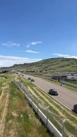 The video shows a race track with three cars driving on it. There is a metal fence along the left side of the track, and grass between the fence and the track. Beyond the track, there is a building and parking lot. In the distance, there is a green, rolling hill. The sky is blue with some clouds. The cars pass by in quick succession, from left to right on the screen, and slightly upward. The red car is the last to drive by.