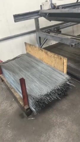 A stack of metal rods rests on wooden planks. A wire cutting machine is partially visible in the background, with a digital control panel displaying numbers. The environment appears to be a workshop or factory floor.