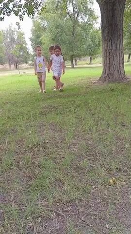 Three young children, two girls and one boy, are walking hand-in-hand across a grassy field. The girl on the left wears a floral print dress and is barefoot. The boy in the middle wears a white t-shirt with a yellow graphic and light-colored shorts, and he is also barefoot. The girl on the right wears a white t-shirt with a pattern of small black dots and matching shorts, and she is wearing white shoes. They are walking towards the right of the frame, with a large tree to their right and other trees and foliage in the background. The ground is covered in green grass and some dry patches. There is a small yellow object, possibly a fallen fruit, on the grass in the lower right corner of the frame.