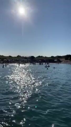 The video shows a sunny, clear day at the beach. The water is a bright blue-green, sparkling under the sunlight. Several people are swimming and playing in the water near the shore. On the beach, there are people, umbrellas, and some vegetation. Across the water, a rocky coastline is visible, with a small building on top. People can also be seen along the coastline. The camera appears to be on a boat or a floating device, gently moving across the water.